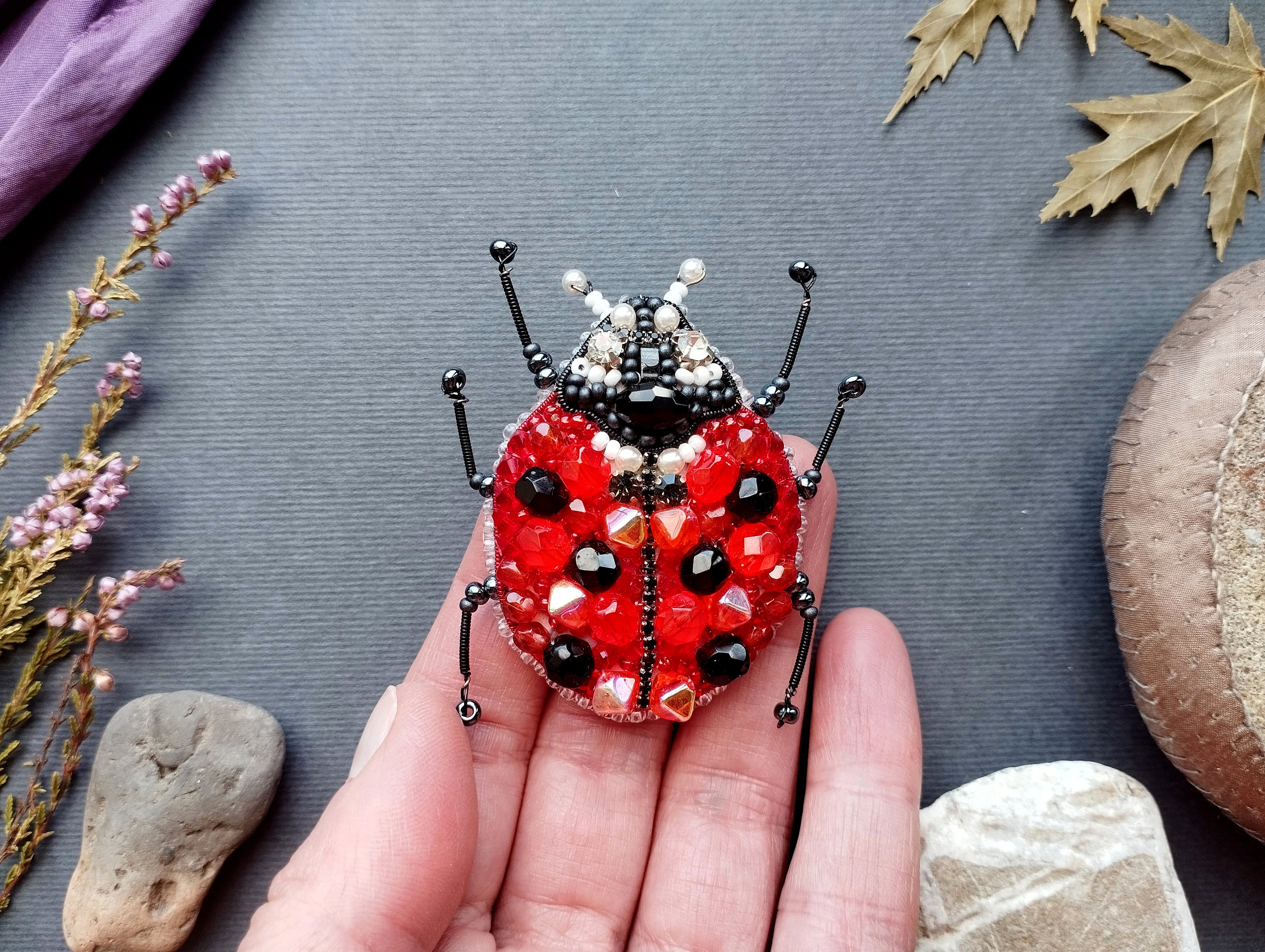A hand is holding a red and black beaded ladybug brooch against a gray background.